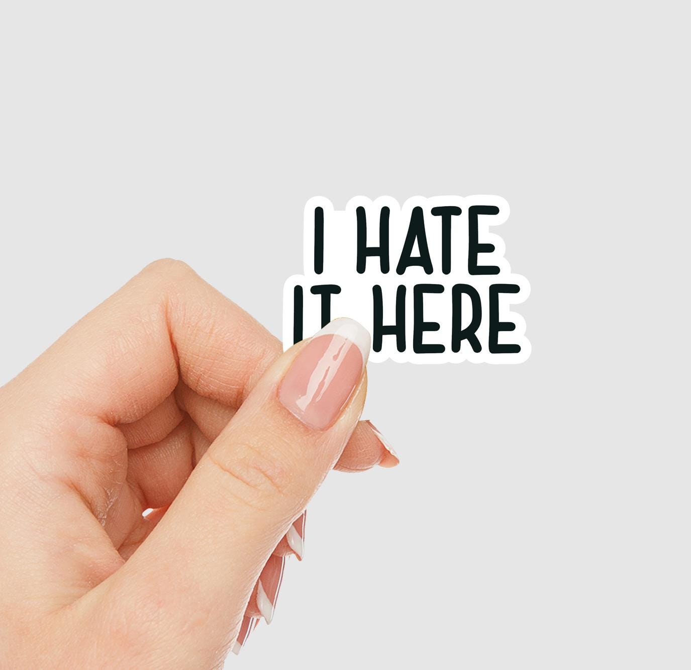 Hand holding a sticker with 'I HATE IT HERE' text on a light gray background