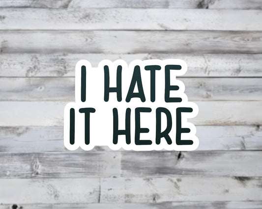 Sticker with 'I HATE IT HERE' text on a wooden background